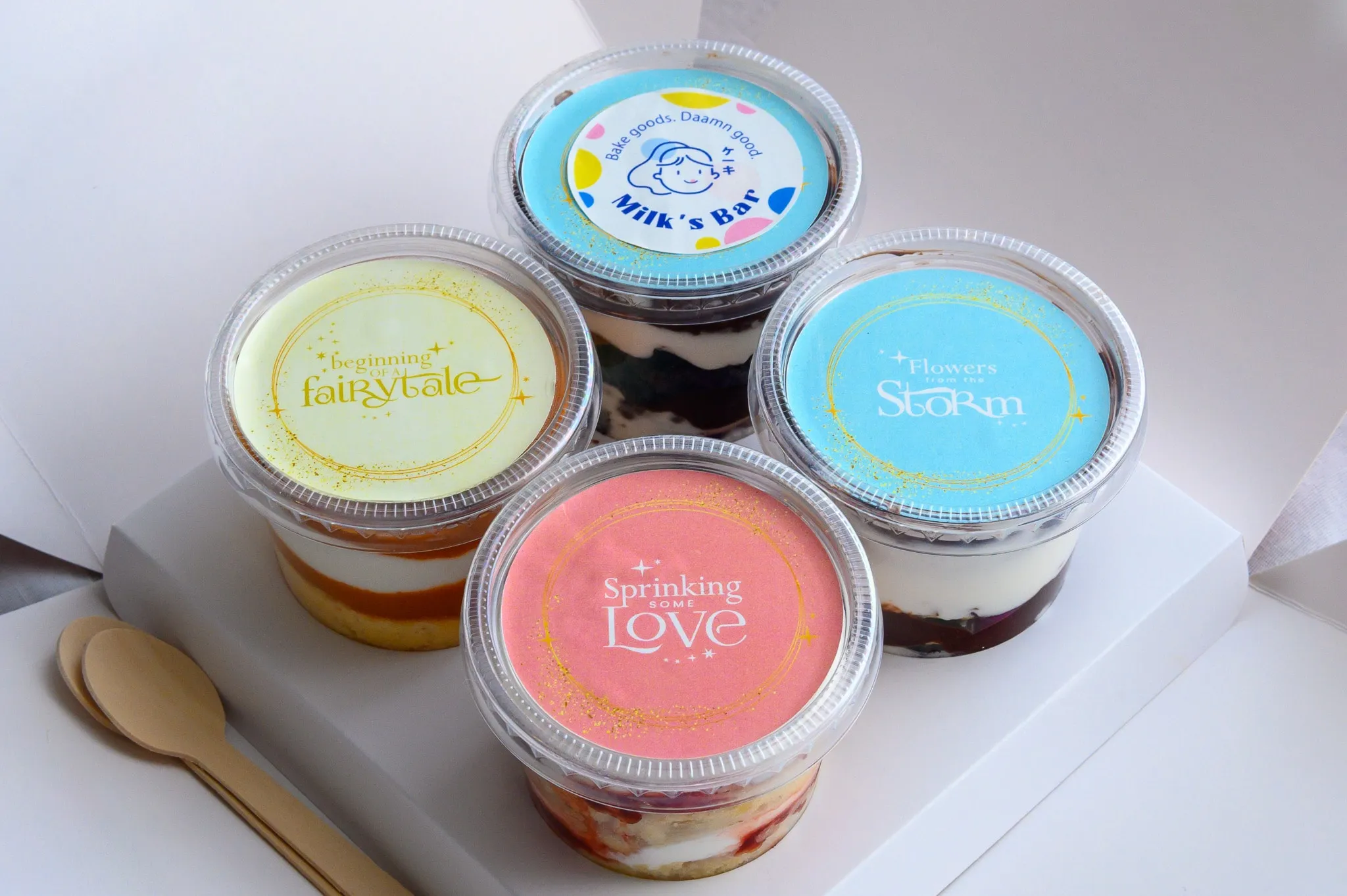 Four layered dessert cups with colorful labeled lids and a wooden spoon