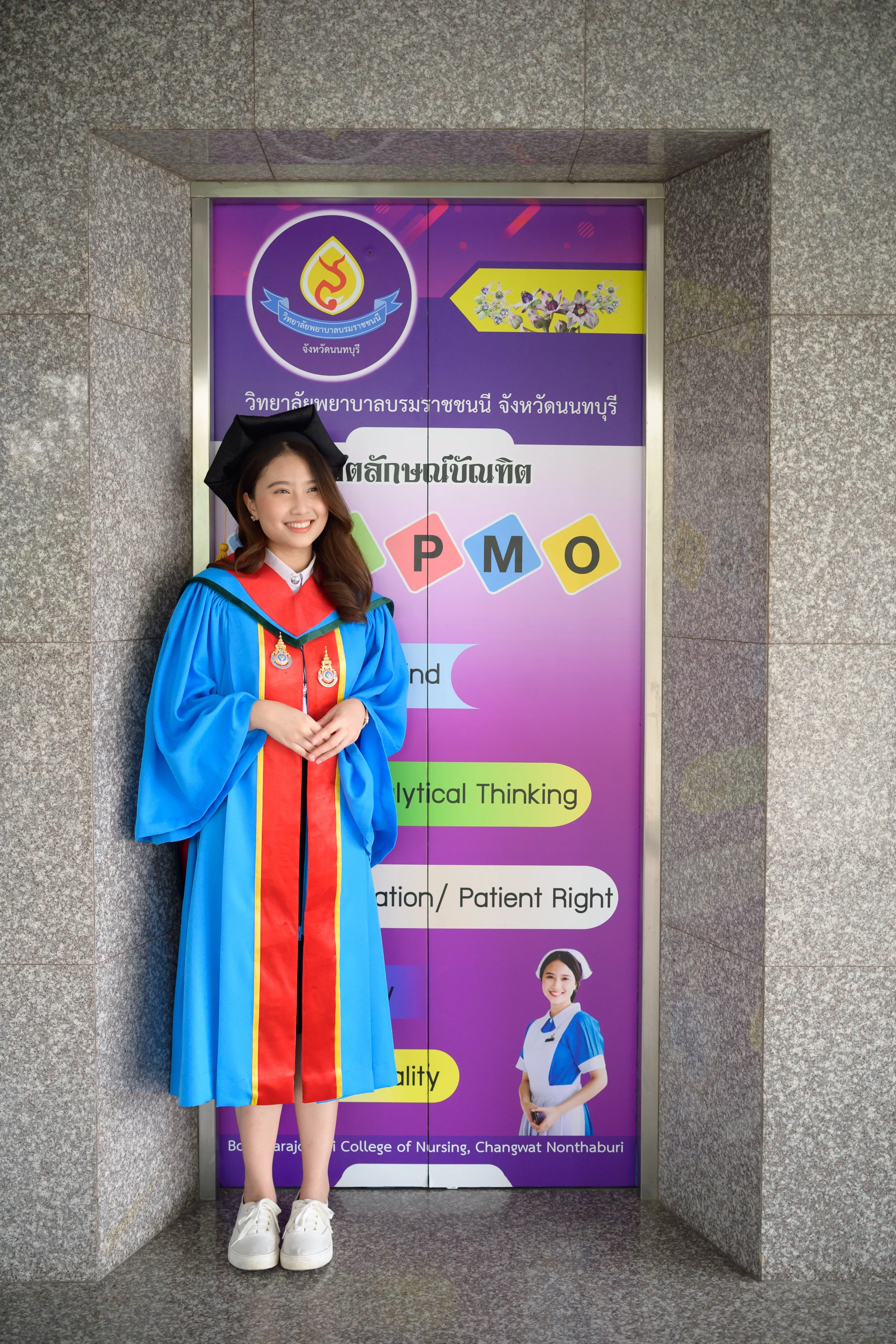 A smiling graduate in a mortarboard and blue gown standing in front of a purple nursing college poster