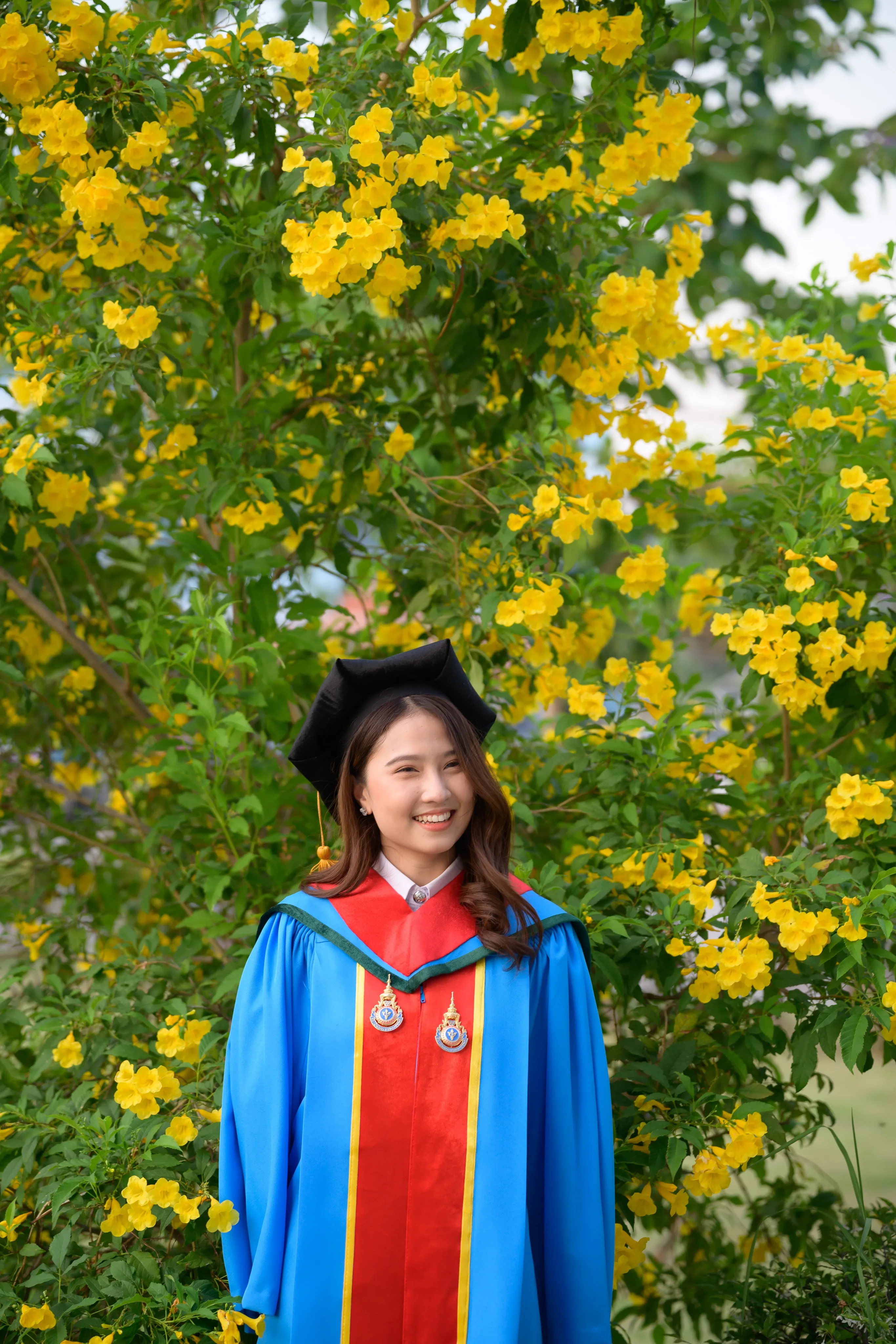 A smiling graduate in a mortarboard and blue gown in front of a bush of yellow flowers
