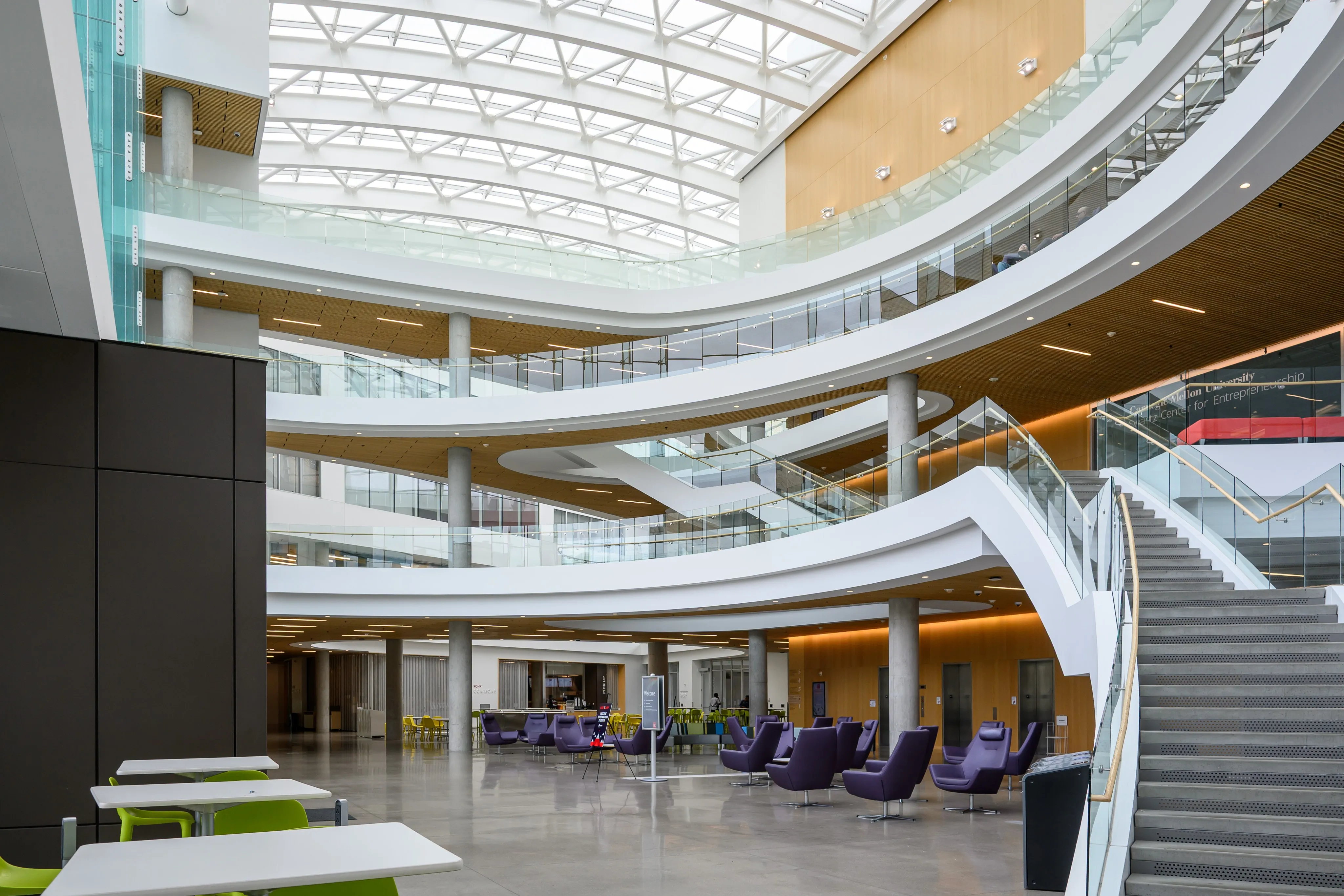 A bright, skylit atrium with sweeping white balconies, a broad staircase, and purple lounge chairs on the ground floor.