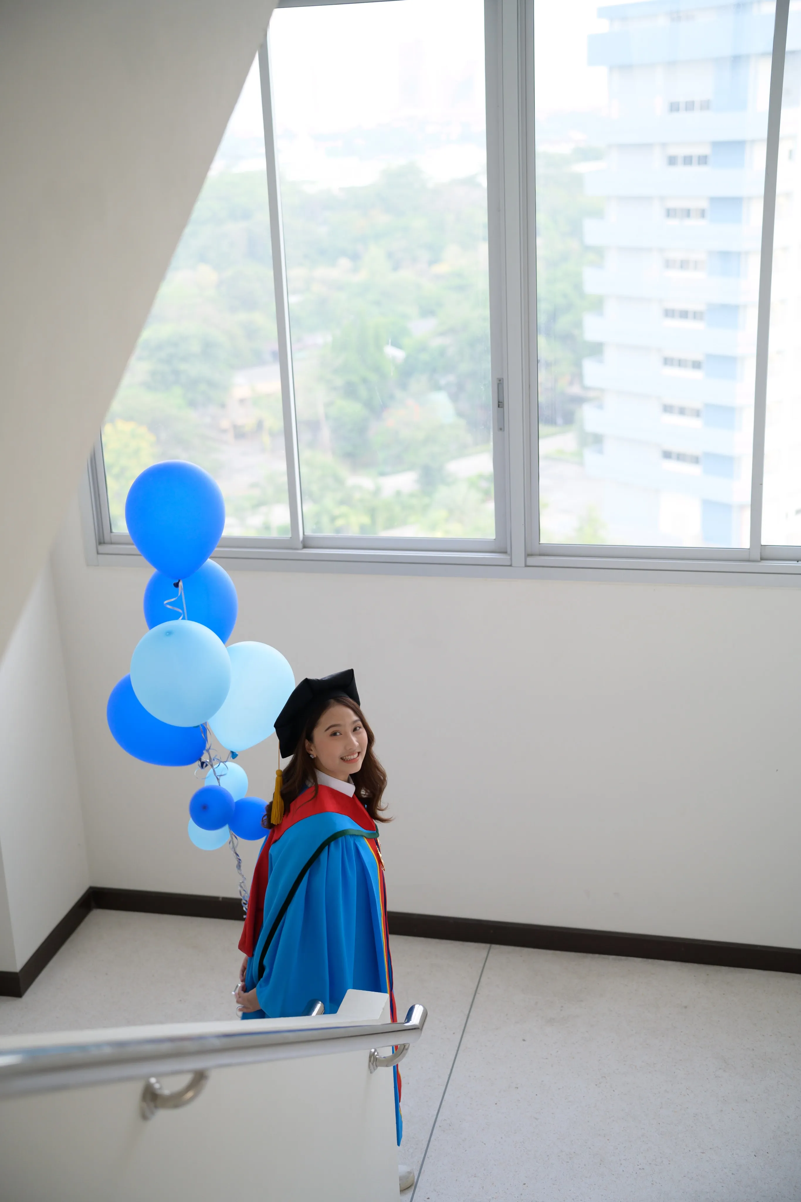 A graduate in a blue gown holding blue balloons in a bright stairwell with city views
