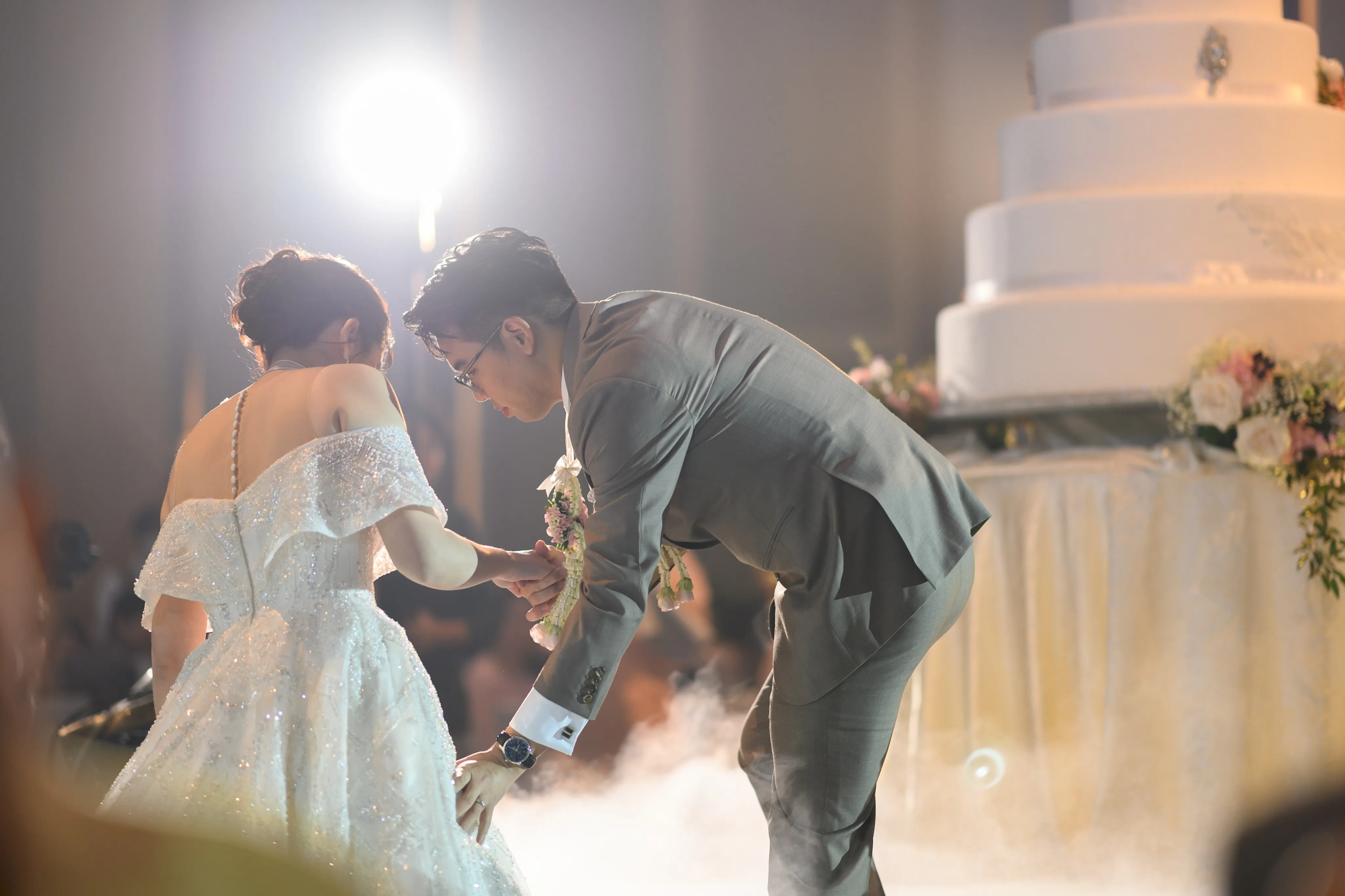 A groom bowing to take the bride's hand beside a tiered white wedding cake at the reception