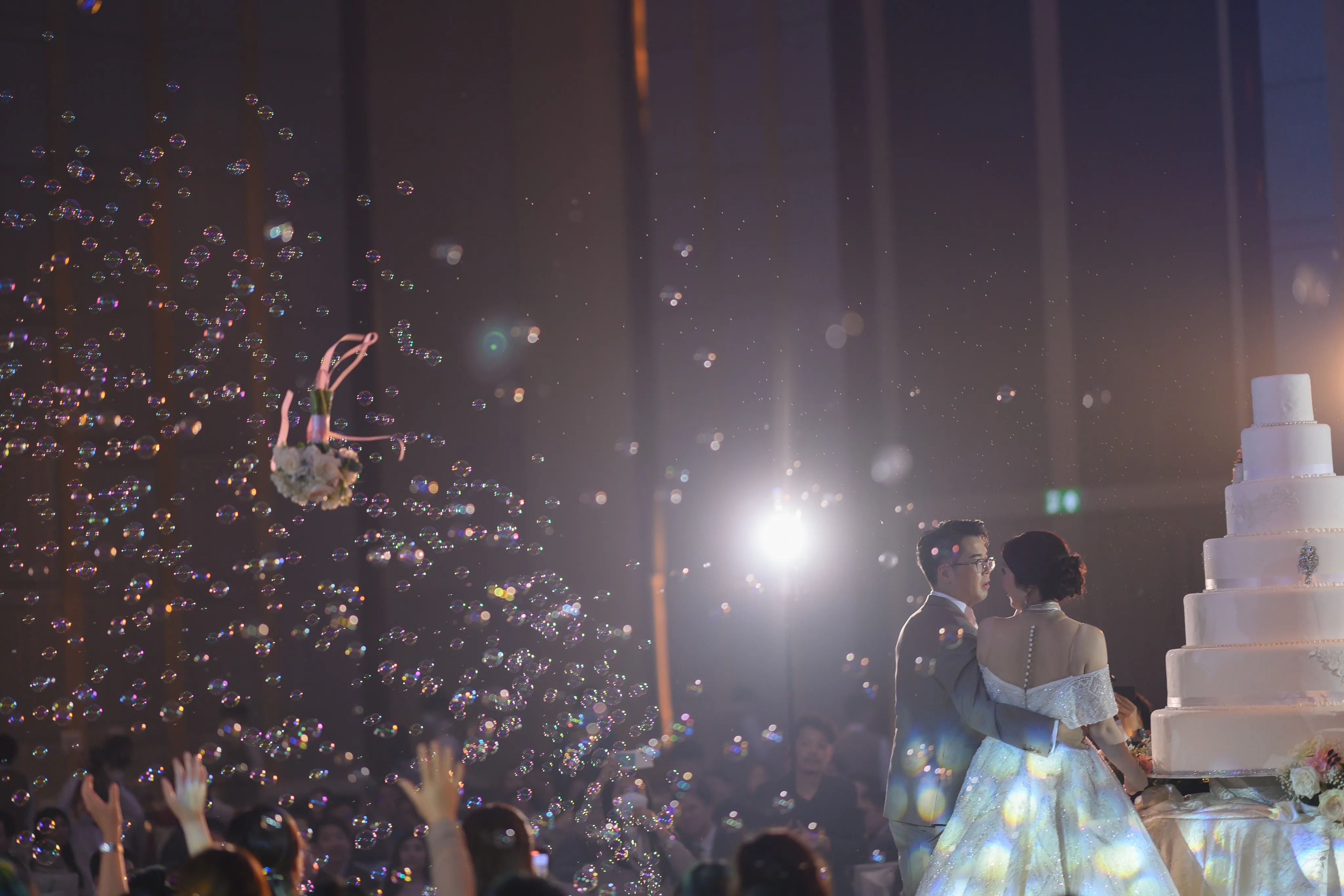 A bride and groom dancing beside a tiered cake as bubbles fill the air above the reception guests