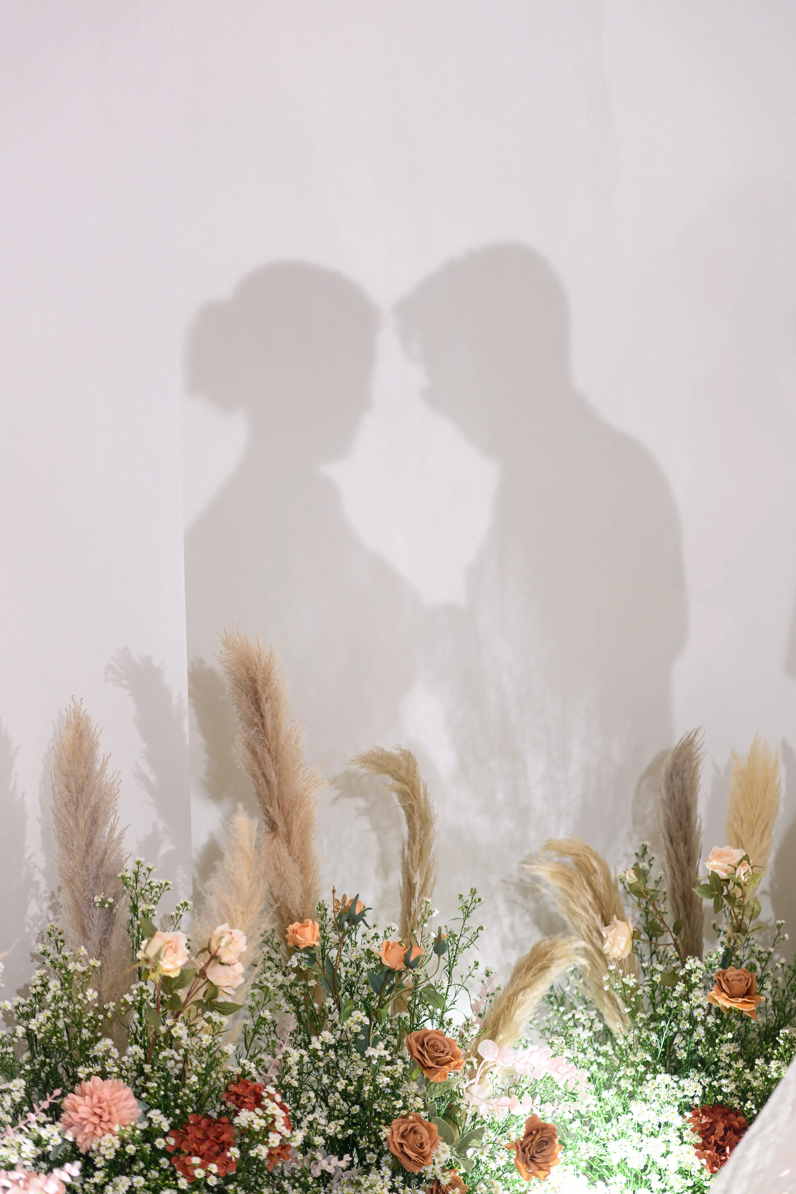 Shadows of a couple touching foreheads on a white wall above pampas grass and flowers