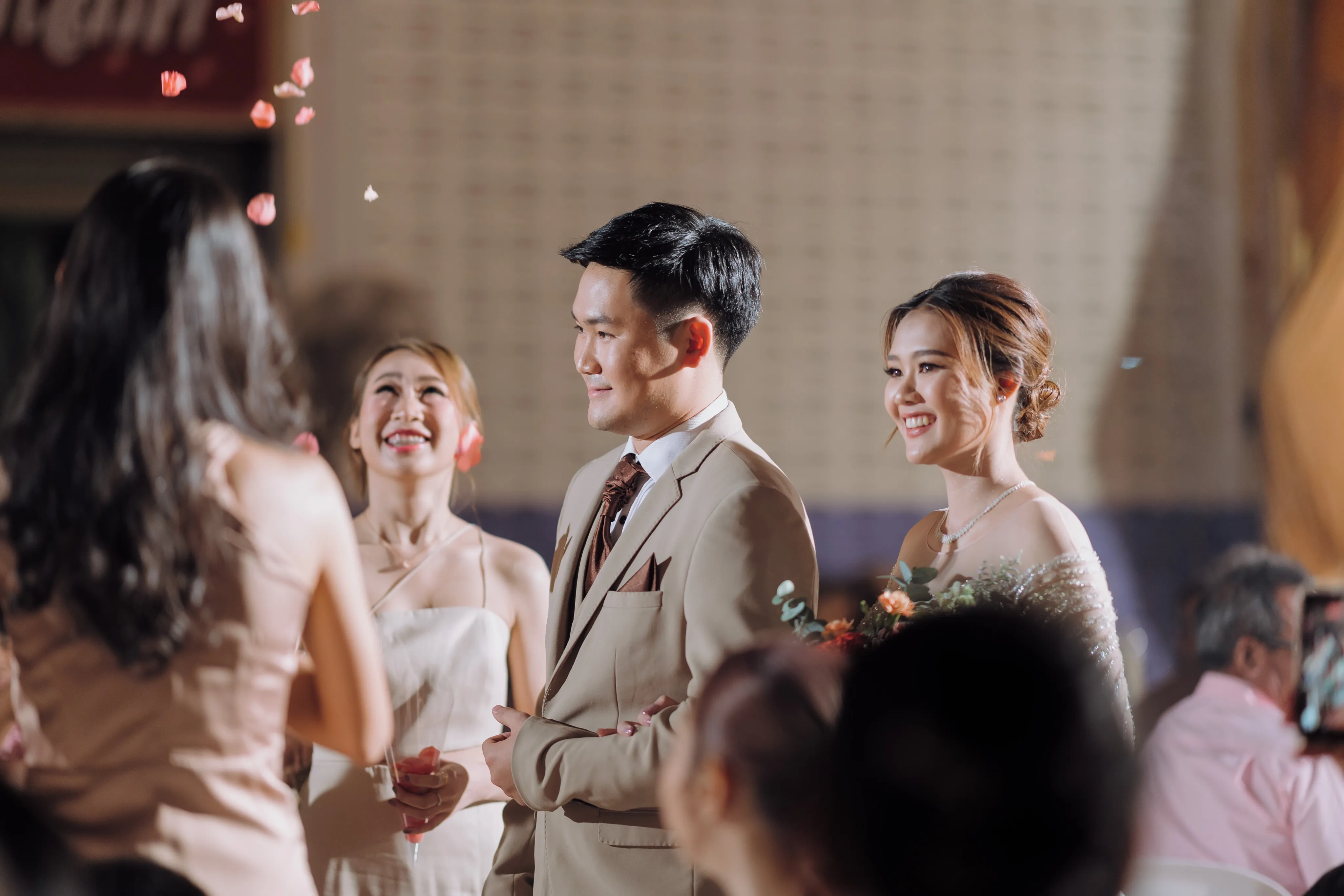 A smiling groom in a beige suit with the bride and a bridesmaid as guests look on