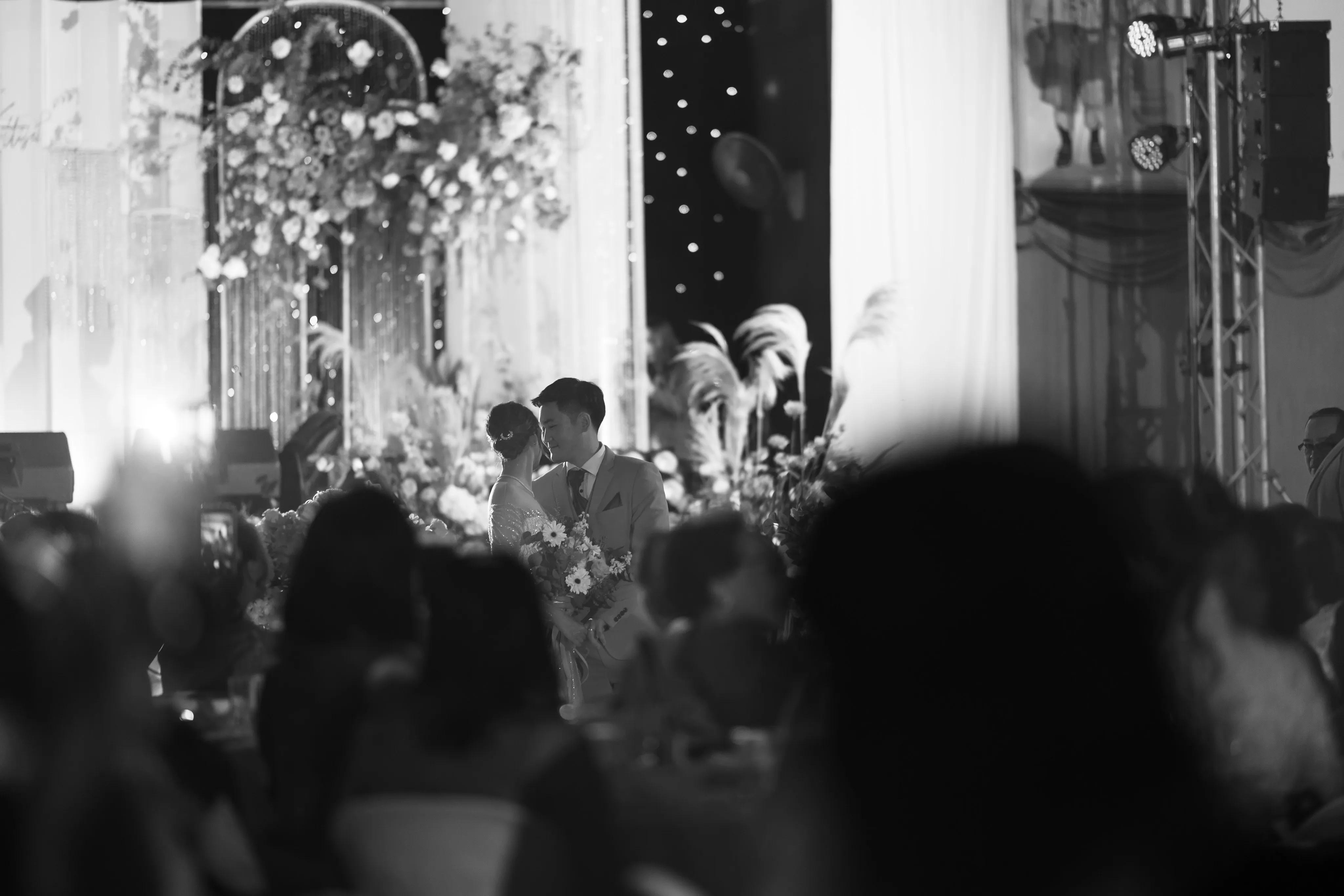 A black-and-white photograph of the bride and groom on stage beyond a crowd of reception guests