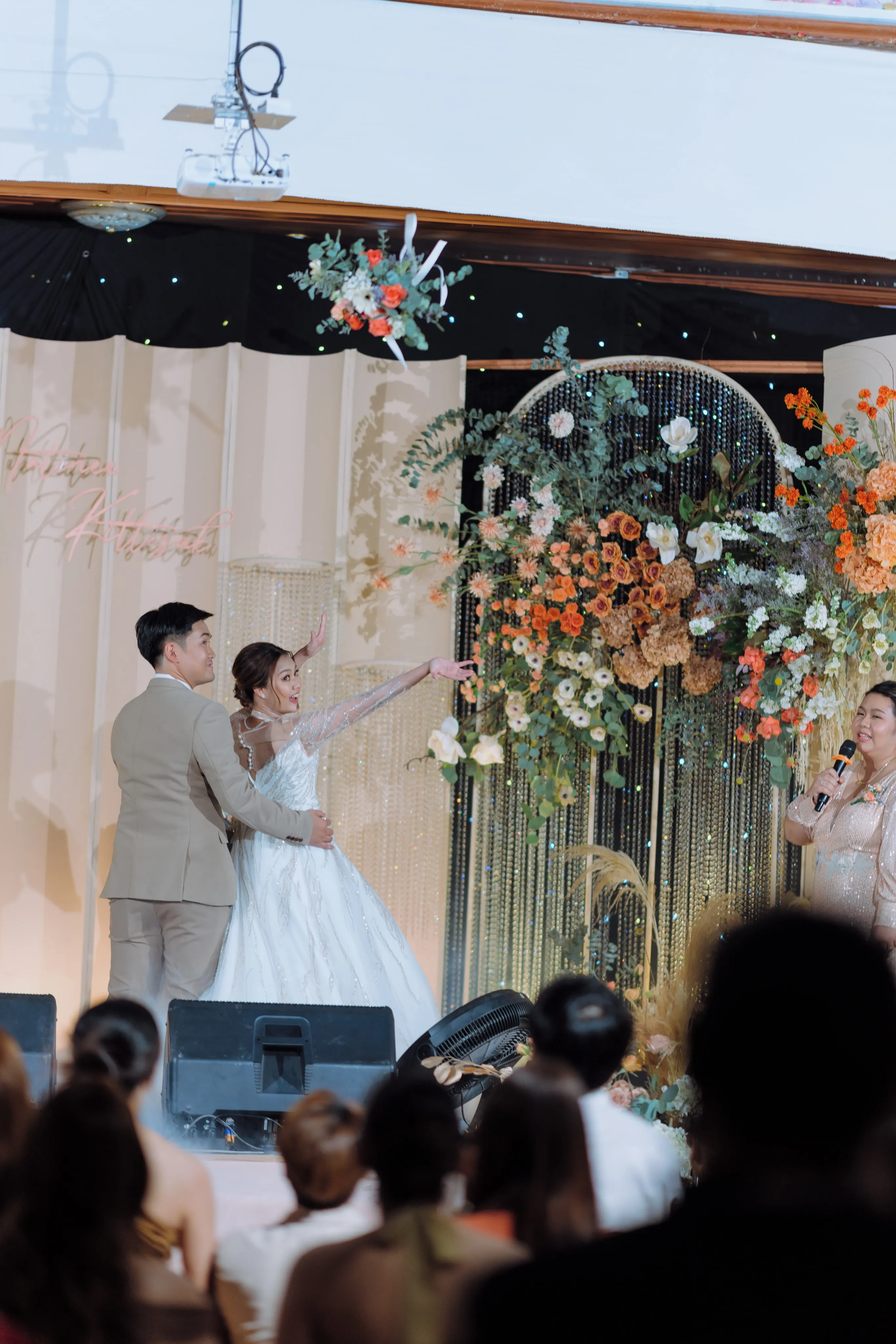 The bride pointing into the crowd beside the groom on a flower-decked stage at the reception