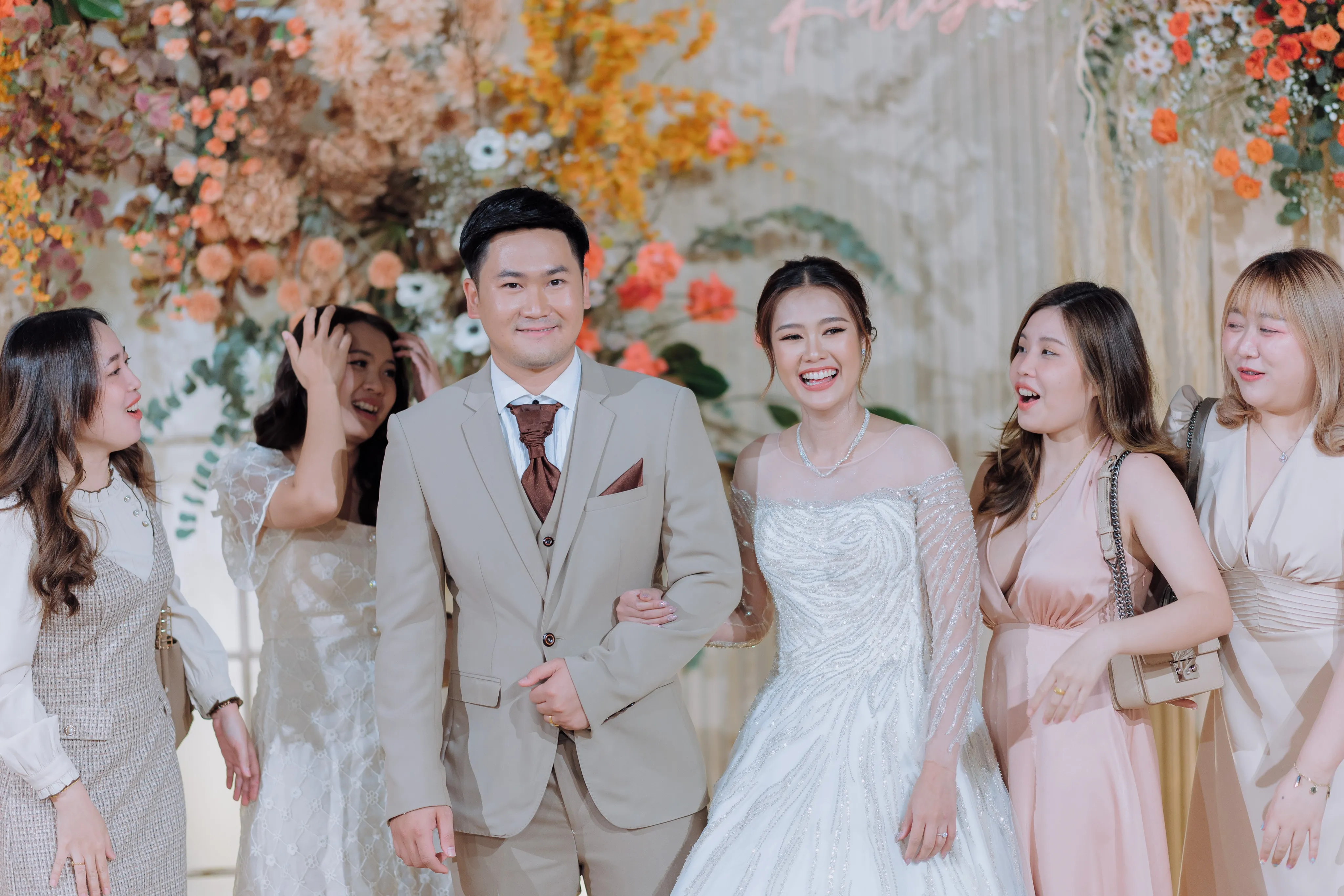 The bride and groom arm in arm with laughing bridesmaids in front of an orange floral backdrop