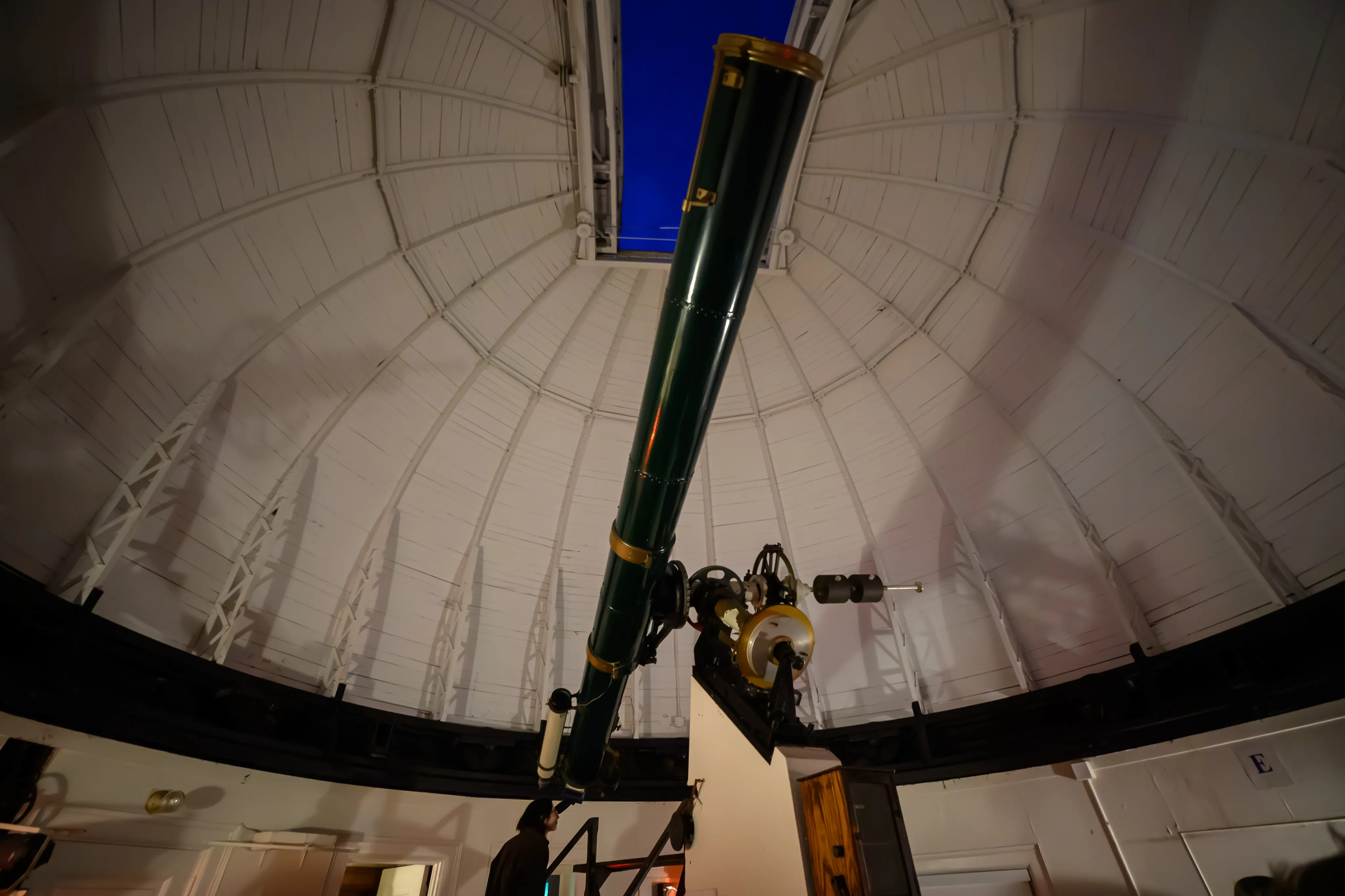 A large green telescope beneath the open dome of Allegheny Observatory, lit against the deep blue evening sky.